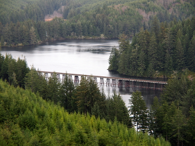 [A raised bridge runs through the foreground arm of the lake while another arm is seen in the back. All edges of the lake are surrounded by tall evergreens.]